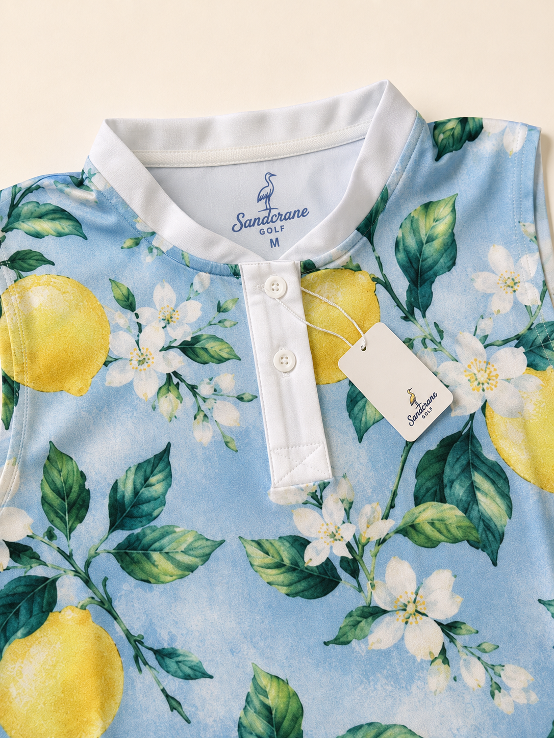 A close-up of a light blue polo shirt with a white collar, featuring a watercolor print of lemons, green leaves, and white flowers. The brand 'Sandtrane Golf' is visible on the inner neck label and the attached hangtag.